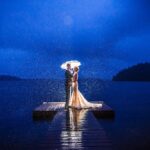 A couple having a destination wedding stands under an umbrella on a dock in the rain.