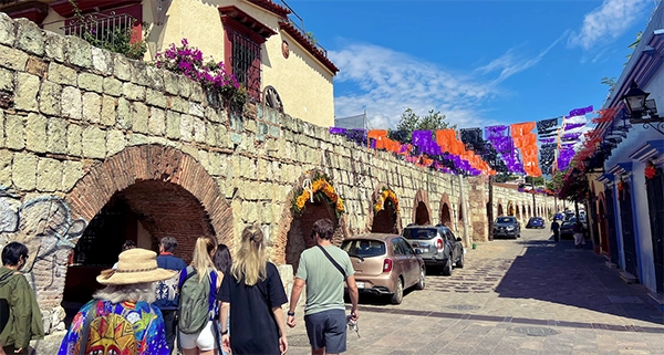 People walk along a cobblestone street lined with stone archways and colorful hanging decorations under a clear blue sky, celebrating the vibrant Day of the Dead in Oaxaca.