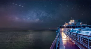A ship deck at night, illuminated under a starry sky and the Milky Way visible. A meteor streaks across the heavens while the deck glistens wet under the ship's lights. The allure of cruises is truly unmatched when accompanied by such celestial wonders.