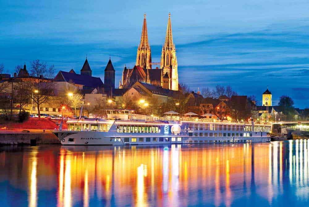 A river cruise boat docked at twilight on the Danube River, with a cathedral and town buildings aglow in the background, their lights shimmering on the water—an enchanting view perfect for those embarking on a Budapest to Munich/Passau journey.