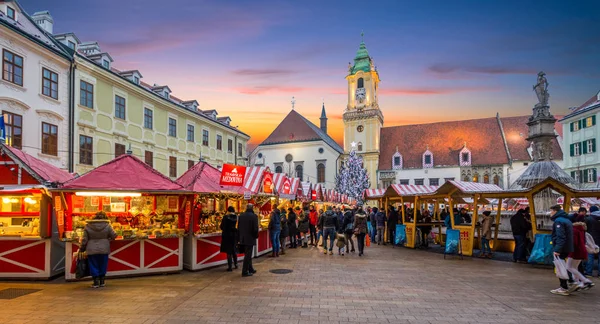 A bustling holiday market at sunset features various stalls with red canopies, surrounded by historic buildings and a church with a tall spire in the background—a perfect stop on a Danube River Cruise from Budapest to Munich.