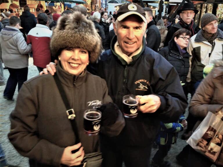 A man and woman wearing winter clothing hold mugs while standing in a bustling holiday market, their excitement palpable as they reminisce about adventures from Budapest to Munich.