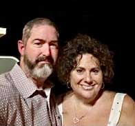A man with a beard and a woman with curly hair smile warmly as they stand together, evoking the joy and charm of exploring holiday markets along a Danube River Cruise against a dark background.